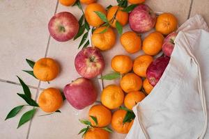 Reife Mandarinen und rote Äpfel in einer wiederverwendbaren Öko-Tasche auf einem Keramikboden. obstlieferung, gesundes essen, vitamine. Draufsicht auf saftige Früchte. foto