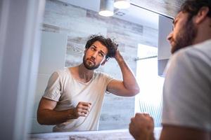 hübscher junger mann, der sein haar mit der hand berührt und sich im badezimmer zu hause pflegt. Weißer metrosexueller Mann, der sich Sorgen um Haarausfall macht und seinen zurückgehenden Haaransatz im Spiegel betrachtet. foto