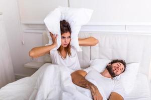 Mann schnarcht, während seine Frau die Ohren mit Kissen bedeckt. Frau leidet darunter, dass ihr männlicher Partner im Bett schnarcht. Geräuschkonzept. echte Menschen. junge gereizte frau, die mit schnarchendem ehemann im bett liegt foto