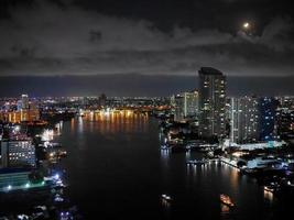 nachtaufnahme, langzeitbelichtung, beleuchtete lichter, hoher winkel, luftaufnahme der skyline, stadtbild, flussufer, chao phraya fluss, mond im hintergrund, bangkok thailand foto