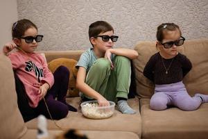 drei kinder sitzen im wohnzimmer, tragen eine 3d-brille und schauen sich filme oder zeichentrickfilme an. foto