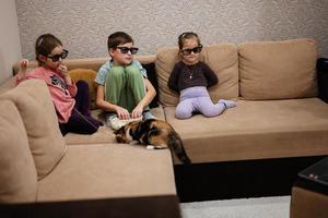drei kinder sitzen im wohnzimmer, tragen eine 3d-brille und schauen sich filme oder zeichentrickfilme an.