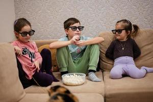 drei kinder sitzen im wohnzimmer, tragen eine 3d-brille und schauen sich filme oder zeichentrickfilme an. foto