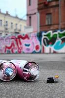 mehrere gebrauchte sprühdosen mit rosa und weißer farbe und kappen zum sprühen von farbe unter druck liegen auf dem asphalt in der nähe der bemalten wand in farbigen graffiti-zeichnungen foto