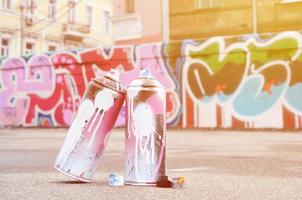 mehrere gebrauchte sprühdosen mit rosa und weißer farbe und kappen zum sprühen von farbe unter druck liegen auf dem asphalt in der nähe der bemalten wand in farbigen graffiti-zeichnungen foto