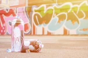 mehrere gebrauchte sprühdosen mit rosa und weißer farbe und kappen zum sprühen von farbe unter druck liegen auf dem asphalt in der nähe der bemalten wand in farbigen graffiti-zeichnungen foto