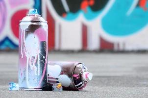 mehrere gebrauchte sprühdosen mit rosa und weißer farbe und kappen zum sprühen von farbe unter druck liegen auf dem asphalt in der nähe der bemalten wand in farbigen graffiti-zeichnungen foto