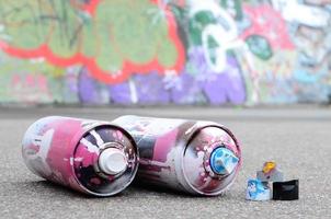 mehrere gebrauchte sprühdosen mit rosa und weißer farbe und kappen zum sprühen von farbe unter druck liegen auf dem asphalt in der nähe der bemalten wand in farbigen graffiti-zeichnungen foto