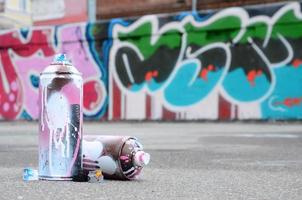 mehrere gebrauchte sprühdosen mit rosa und weißer farbe und kappen zum sprühen von farbe unter druck liegen auf dem asphalt in der nähe der bemalten wand in farbigen graffiti-zeichnungen foto