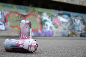 ein paar gebrauchte sprühdosen mit rosa und weißer farbe liegen auf dem asphalt vor dem hintergrund einer bemalten wand in bunten graffiti-zeichnungen foto