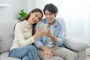 asiatisches Paar lächelt und nutzt Smartphone glücklich zum Online-Shopping. Mann bereit, Geburtstagsgeschenk für schöne Frau zu bezahlen. Unterkunft online finden für Flitterwochen, App online, Flugtickets buchen. foto
