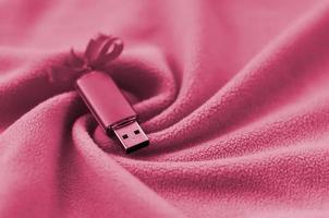 USB-Flash-Speicherkarte mit Schleife liegt auf einer Decke aus weichem und pelzigem Fleece-Stoffbild, getönt in Viva Magenta, Farbe des Jahres 2023 foto