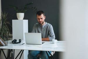 junger lächelnder geschäftsmann im lässigen grauen langarm, der am laptop im dunklen modernen büroraum arbeitet foto