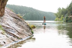 die schlanke junge frau im grünen sweemsuit auf dem sup-boot mit ruder, das auf dem fluss schwimmt, wochenendausflug und lokale reise foto