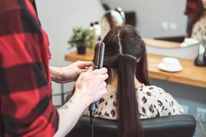 Friseurfriseur, der langes Haar mit Haareisen für schöne junge Frau im Arbeitsmoment des Schönheitssalons anredet foto