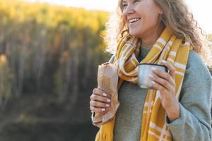 junge glückliche schöne reisende mit lockigem haar, die hotdog isst und tee auf dem hintergrund der berge und des flusses trinkt und auf der herbstnatur wandert foto