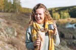 junge glückliche schöne reisende mit lockigem haar, die hotdog isst und tee auf dem hintergrund der berge und des flusses trinkt und auf der herbstnatur wandert foto