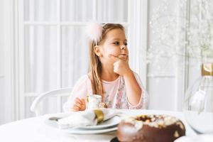 lustiges süßes kleines mädchen mit langen haaren in hellrosa kleid mit schokoladenkuchen in den händen auf festlichem tisch im hellen wohnzimmer zu hause. Weihnachtszeit, Geburtstagskind foto