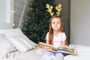 süßes kleines Mädchen in rosafarbenem Kleid liest ein Buch, das auf dem Bett im Zimmer mit Weihnachtsbaum sitzt. Kind in der Felge mit Hirschhörnern im Landhaus in der Weihnachtszeit, frohes neues Jahr foto