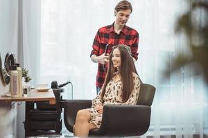 Friseurfriseur, der langes Haar mit Haareisen für schöne junge Frau im Arbeitsmoment des Schönheitssalons anredet foto