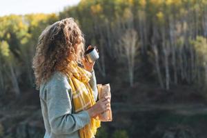 junge glückliche schöne reisende mit lockigem haar, die hotdog isst und tee auf dem hintergrund der berge und des flusses trinkt und auf der herbstnatur wandert foto