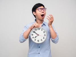 Der junge asiatische Mann mit Brille, der eine Uhr hält und gähnt, fühlt sich schläfrig und isoliert foto