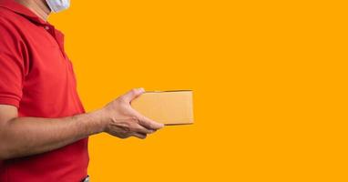 Seitenansicht. Zusteller, der eine chirurgische Maske und medizinische Handschuhe in roter Uniform trägt, isoliert auf gelbem Hintergrund, hält Paketboxen für den Versand von Paketen per Post. Tapete Leinwand breit. foto