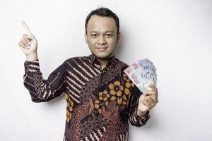 ein glücklicher junger asiatischer mann trägt ein batikhemd und zeigt auf den kopierraum auf ihm, während er geld in indonesischer rupiah hält, das durch weißen hintergrund isoliert wird foto