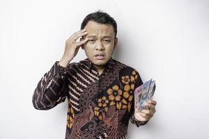 ein gestresster asiatischer mann, der ein batikhemd trägt und geld in indonesischer rupiah hält, das durch weißen hintergrund isoliert wird, sieht deprimiert aus foto