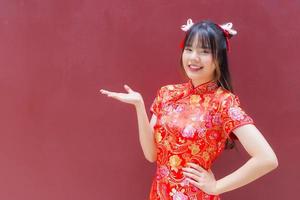 süßes asiatisches mädchen mit langen haaren, das ein rotes cheongsam-kleid im chinesischen neujahrsthema trägt, während sie ihre hand zeigt, um etwas auf rotem hintergrund zu präsentieren. foto