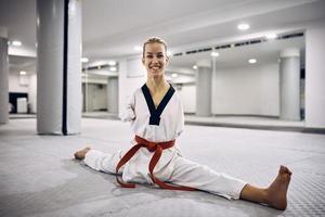 glückliche sportliche Frau ohne die oberen Gliedmaßen, die sich auf dem Boden strecken, während sie Taekwondo üben und in die Kamera schauen. foto