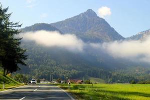 reise nach sankt-wolfgang, österreich. die straße zwischen grünen wiesen mit den bergen in den wolken im hintergrund am sonnigen tag. foto