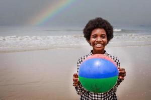 Porträt eines glücklichen afroamerikanischen Jungen, der Wasserball an einem tropischen Strand hält. ethnisch vielseitig foto