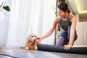 Junge asiatische Frau rollt schwarze Yogamatte aus, um mit ihrem Hund zu Hause Yoga zu spielen foto