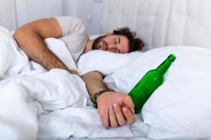 nach Partytrinken. betrunkener bärtiger erwachsener mann, der auf dem bett liegt und schläft, nachdem er viel alkohol getrunken hat, leere flasche auf dem bett, alkoholismus, alkoholsucht und menschenkonzept foto