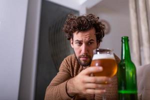 Nach der Arbeit sitzt ein depressiver Typ auf dem Sofa und trinkt ein kaltes Bier. Hand hält ein Glas, Mann trinkt allein zu Hause. alkoholismus, alkoholsucht und menschenkonzept foto