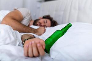 nach dem Trinken. betrunkener bärtiger erwachsener mann, der auf dem bett liegt und schläft, nachdem er viel alkohol getrunken hat, leere flasche auf dem bett, alkoholismus, alkoholsucht und menschenkonzept foto