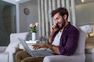 lächelnder gutaussehender mann, der auf dem smartphone spricht und einen laptop benutzt, während er zu hause sitzt. freiberufliche Karriere. kerl, der auf dem handy mit dem laptop spricht, der zu hause auf dem boden sitzt. Freiraum für Text foto