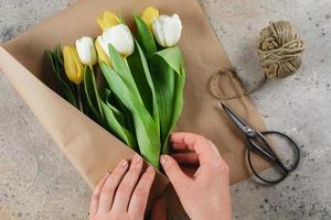 Weiße und gelbe Tulpen in einem Bastelpaket. weibliche hände, die einen blumenstrauß in kraftpapier arrangieren. Frühlingsferienkonzept