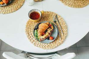 frisches croissant-sandwich mit schinken, rührei, tomate mit tee auf dem tisch, draufsicht foto