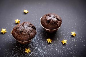 Zwei köstliche Schokoladenmuffins, umgeben von goldenen Sternen und weißem Pulver auf schwarzem Papierhintergrund foto