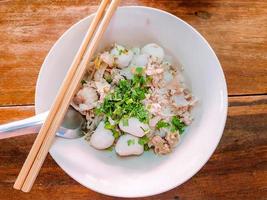 Draufsicht und Nahaufnahme von leckeren chinesischen Nudeln ohne Suppe mit Schweinefleischbällchen und Stück grobem Schweinefleisch in weißer Keramikschale mit Essstäbchen und Löffel auf Holztischhintergrund. foto