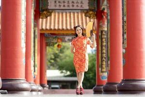 asiatische frau im roten cheongsam qipao kleid, das papierfächer hält, während sie den chinesischen buddhistischen tempel während des neuen mondjahres für traditionelles kulturkonzept besucht foto