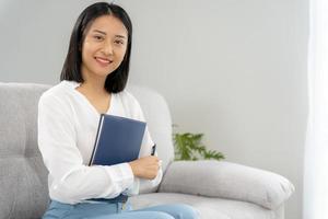 asiatische geschäftsfrau, die ein buch im büro lächelt und hält. schöne und gut aussehende asiatin sitzt auf dem sofa. weibliche Porträts. foto