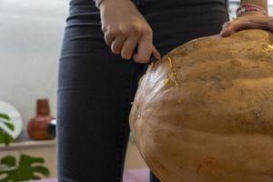 Frau, die großen orangefarbenen Kürbis für Halloween schnitzt, während sie zu Hause am Holztisch sitzt foto