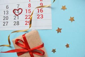 Kalenderseite mit Herz in Rot am 14. Februar des Valentinstags, Geschenkbox auf blauem Hintergrund. Draufsicht mit Platz für Grüße. Konzept des Valentinstags, Feiertag foto