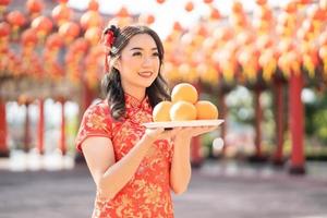 frohes chinesisches neujahr. schöne asiatische frau, die traditionelles cheongsam qipao kleid trägt, das frische orangen im chinesischen buddhistischen tempel hält. Emotion Lächeln foto
