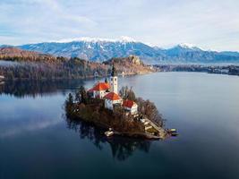 luftdrohnenansicht der sonnenaufgangswinterlandschaft des magischen sees bled in slowenien. ein Wintermärchen für romantische Erlebnisse. Berge mit Schnee im Hintergrund. Kirche der Gottesmutter im See. foto