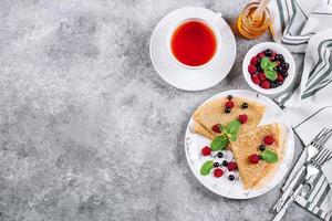 Köstliches Crêpes-Frühstück auf grauem Betontischhintergrund. orthodoxer Feiertag Masleniza. Pfannkuchen mit schwarzen Johannisbeeren, Himbeeren, Glas Honig und Minze. weiße Tasse Tee. Draufsicht, Kopierbereich foto