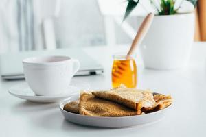 leckeres Frühstück. Weißer Desktop-Tisch mit Laptop und frischen Crêpes-Pfannkuchen, Tasse Tee und Honigglas. Freelancer-Konzept. gesundes frühstück essen. foto